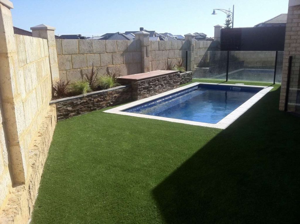 Backyard featuring a small swimming pool surrounded by artificial grass and stone walls.